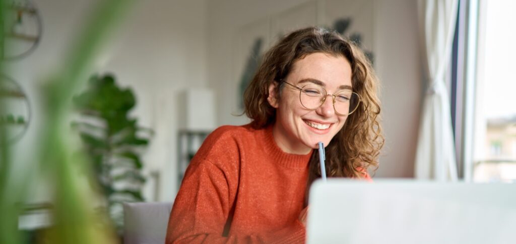 Happy,Young,Woman,Using,Laptop,Sitting,At,Desk,Writing,Notes